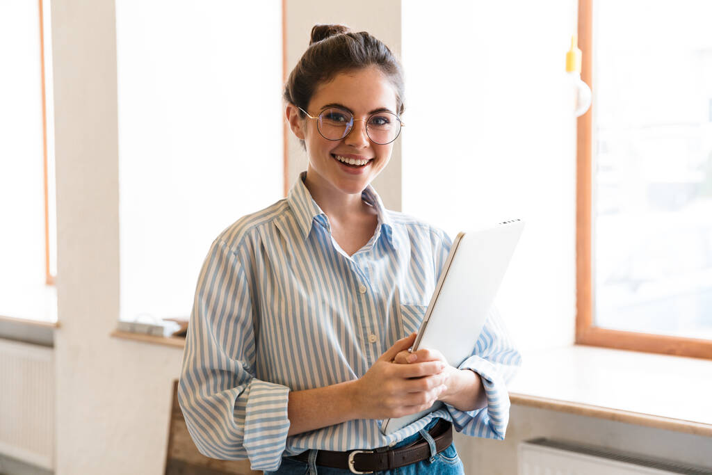 Usmívající se mladá atraktivní brunetka podnikatelka nesoucí notebook, zatímco stojí u okna uvnitř - Fotografie, Obrázek