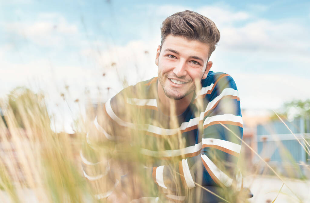 Krásný chlap s bílými zuby, usmívající se kamerou přes rozmazanou trávu, selektivní zaostření. Venkovní - venkovní - Fotografie, Obrázek