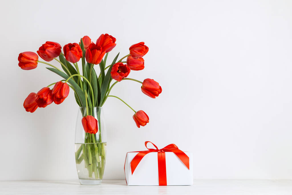 Tulipes rouges dans un vase et cadeau sur la table, sur fond blanc. Carte postale vierge
. - Photo, image