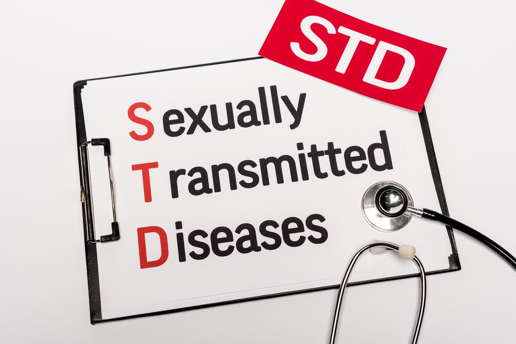 bovenaanzicht van klembord met seksueel overdraagbare aandoeningen in de buurt van papier met STD-letters en stethoscoop geïsoleerd op wit - Foto, afbeelding