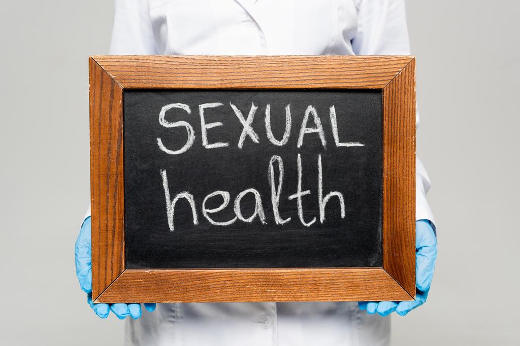 bijgesneden weergave van arts in witte jas en latex handschoenen met krijtbord met seksuele gezondheid belettering geïsoleerd op grijs  - Foto, afbeelding
