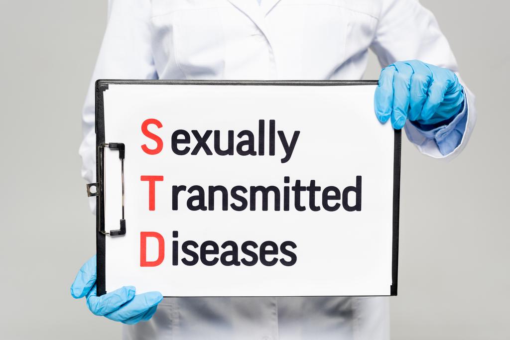 bijgesneden zicht van arts in latex handschoenen met schoolbord met seksueel overdraagbare aandoeningen belettering geïsoleerd op grijs  - Foto, afbeelding