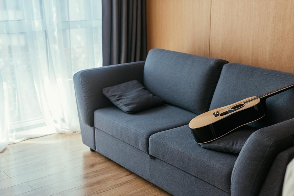interieur van woonkamer met akoestische gitaar op grijze bank en zonlicht in groot raam - Foto, afbeelding