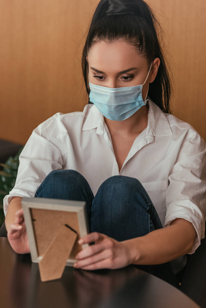 sad young woman in protective mask sitting at table and looking at photo frame - Photo, Image
