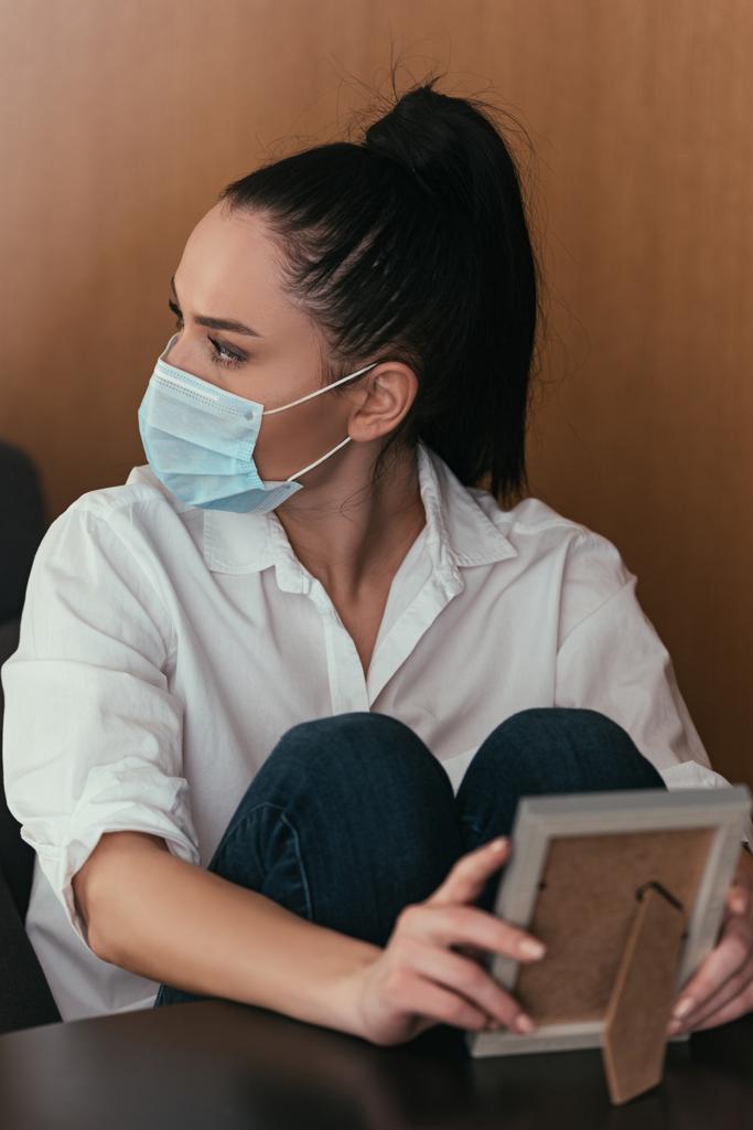 καταθλιπτική νεαρή γυναίκα με ιατρική μάσκα κοιτάζει αλλού ενώ κρατά το πλαίσιο φωτογραφιών - Φωτογραφία, εικόνα