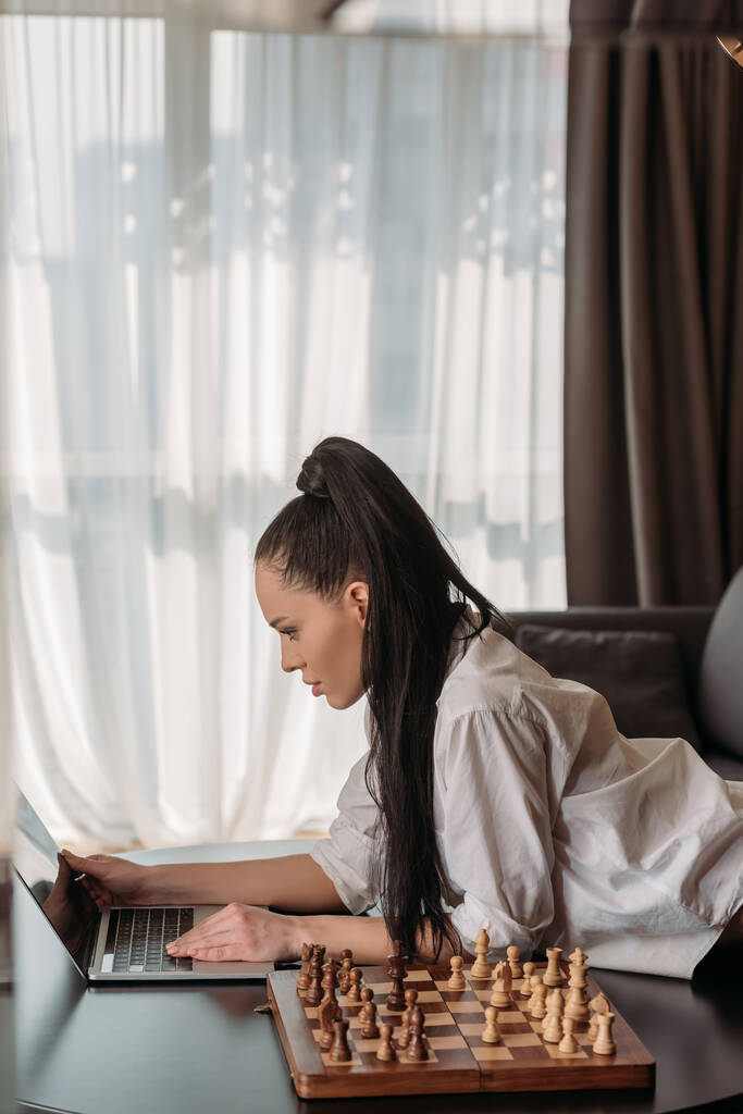 Çekici bir kadının masadaki satranç tahtası yanında boş ekranla dizüstü bilgisayara dokunuşunun yan görüntüsü - Fotoğraf, Görsel