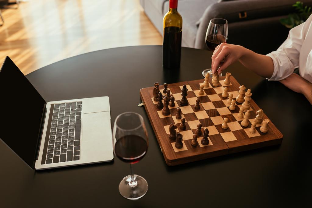 καλλιεργημένη άποψη της γυναίκας που παίζει σκάκι κοντά στο laptop με οθόνη blabk και κόκκινο κρασί στο τραπέζι - Φωτογραφία, εικόνα