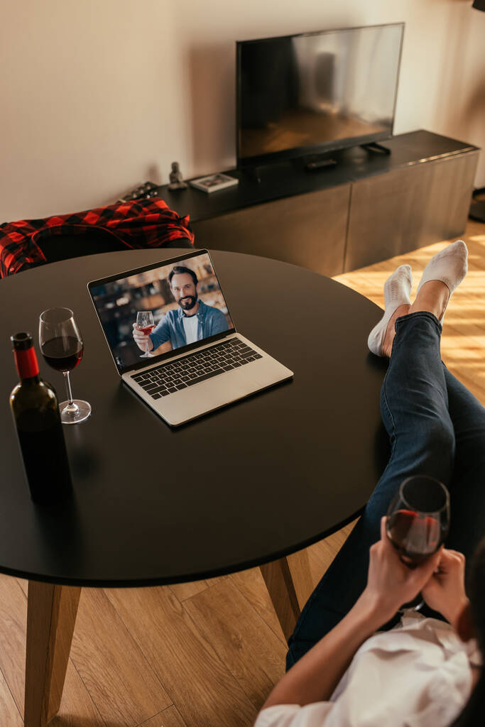 赤ワインのグラスを持っているラップトップの画面上の女性と若い男のクロップドビュー - 写真・画像