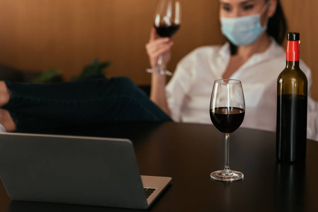 επιλεκτική εστίαση της γυναίκας σε προστατευτική μάσκα κρατώντας ποτήρι κόκκινο κρασί κατά τη διάρκεια της συνομιλίας βίντεο στο laptop - Φωτογραφία, εικόνα