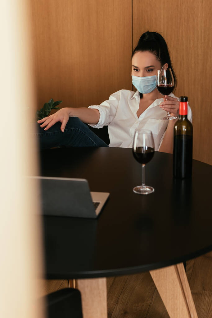 fiatal nő védő maszkban kezében üveg vörösbor közben video chat laptop - Fotó, kép