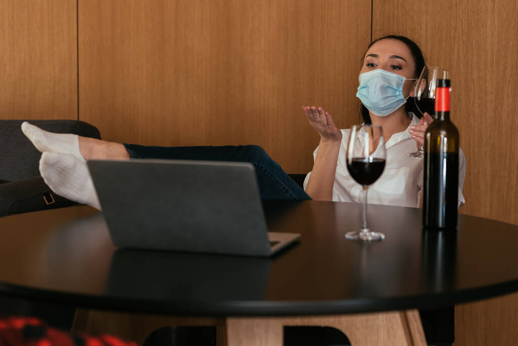 mujer joven en máscara médica el envío de beso de aire durante el chat de vídeo cerca de la botella y vasos de vino tinto
 - Foto, Imagen