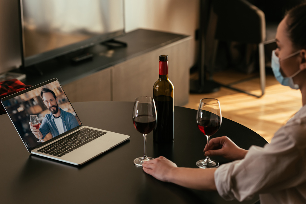 selektivní zaměření mladé ženy v lékařské masce s video chat s přítelem v blízkosti brýle a láhev červeného vína - Fotografie, Obrázek