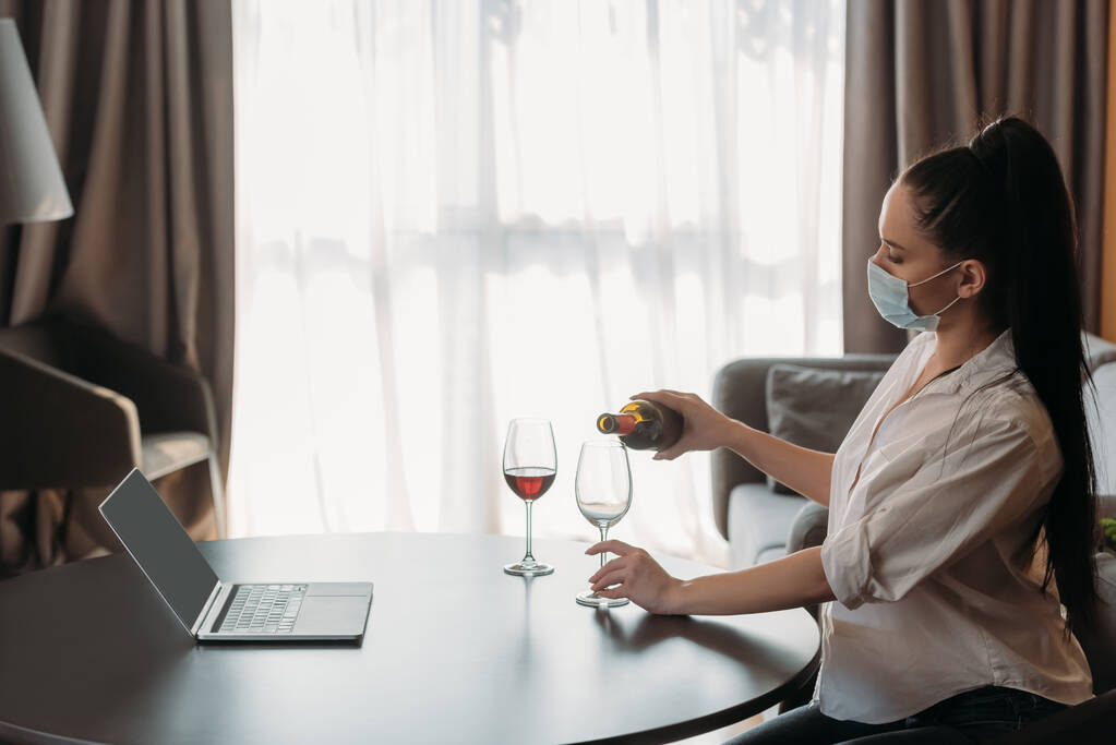 Koruyucu maskeli genç bir kadının, boş ekranlı dizüstü bilgisayarın yanındaki bardağa şarap dolduruşunun yan görüntüsü. - Fotoğraf, Görsel