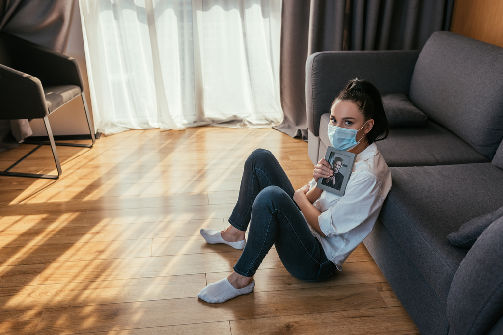 depresja dziewczyna w medycznej masce siedzi na podłodze ze zdjęciem chłopaka i patrząc w aparat - Zdjęcie, obraz