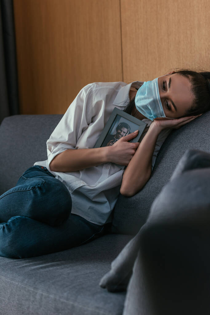 καταθλιπτική νεαρή γυναίκα με ιατρική μάσκα κρατώντας φωτογραφία του φίλου της κοντά στο στήθος, ενώ κάθεται στον καναπέ - Φωτογραφία, εικόνα