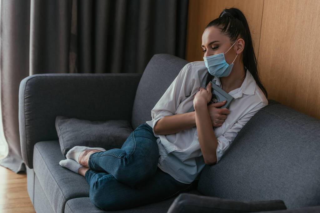 mujer joven estresada en máscara médica sosteniendo el marco de la foto cerca del pecho mientras está sentado en el sofá
 - Foto, Imagen
