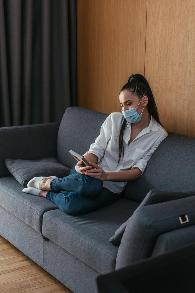 депрессивная молодая женщина в медицинской маске смотрит на фото рамку, сидя на диване
 - Фото, изображение