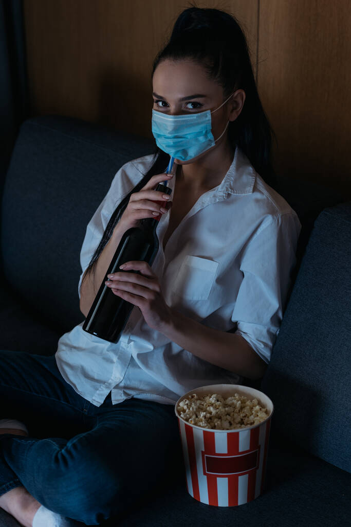 femme déprimée dans un masque médical tenant une bouteille de vin et regardant la caméra près du seau de maïs soufflé
 - Photo, image