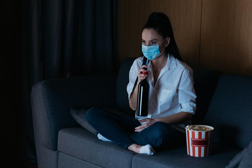 depressieve vrouw in medisch masker tv kijken terwijl zitten op de bank in de buurt van popcorn emmer en het houden van fles wijn  - Foto, afbeelding
