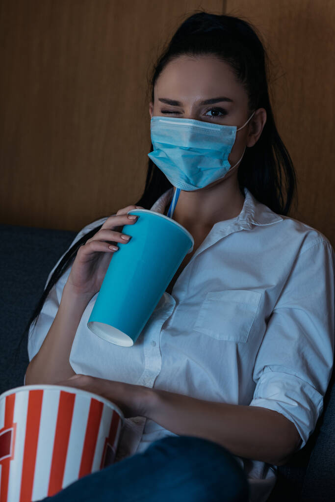 jonge vrouw in medisch masker knipperen op camera terwijl het vasthouden van frisdrank en popcorn  - Foto, afbeelding