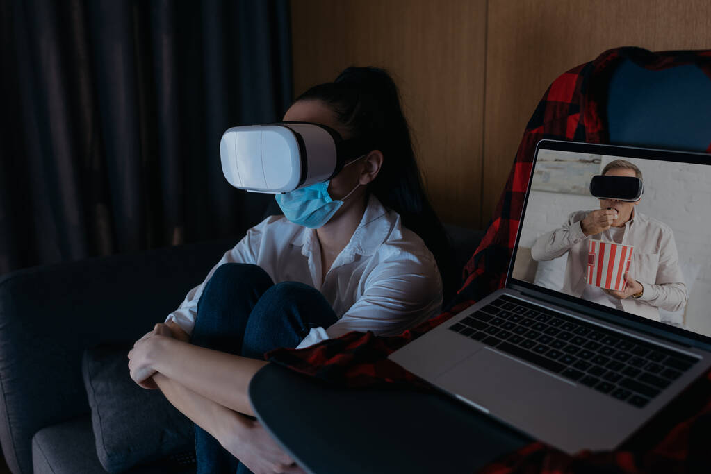 młoda kobieta siedzi na kanapie w pobliżu laptopa z chłopakiem jedząc popcorn za pomocą zestawów słuchawkowych vr razem - Zdjęcie, obraz