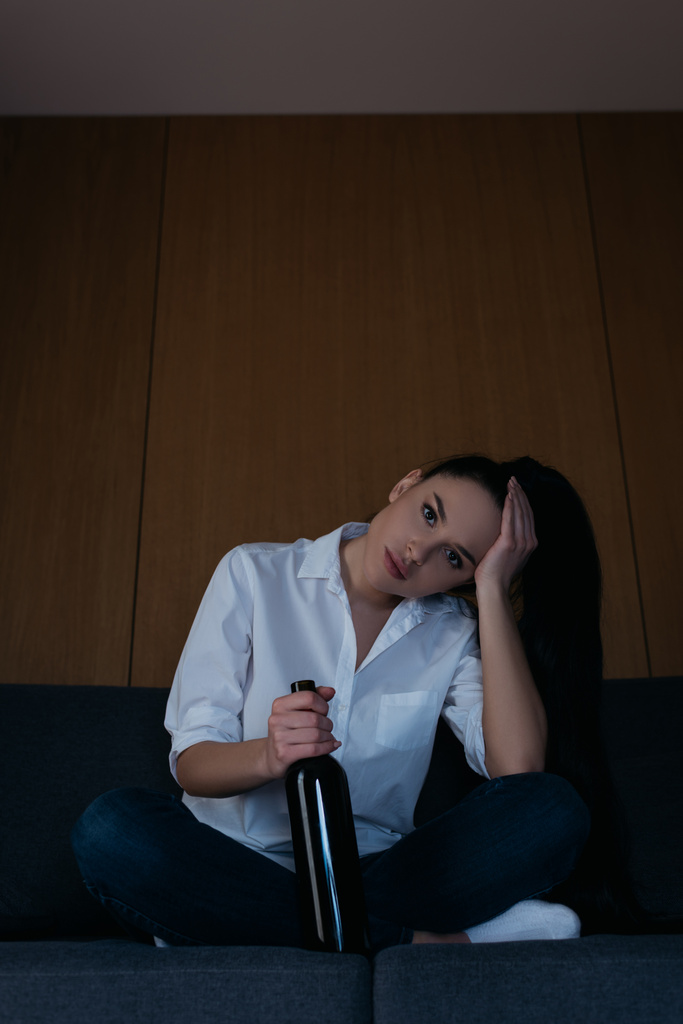 depressed woman touching head and looking at camera while sitting on sofa with bottle of wine - Photo, Image
