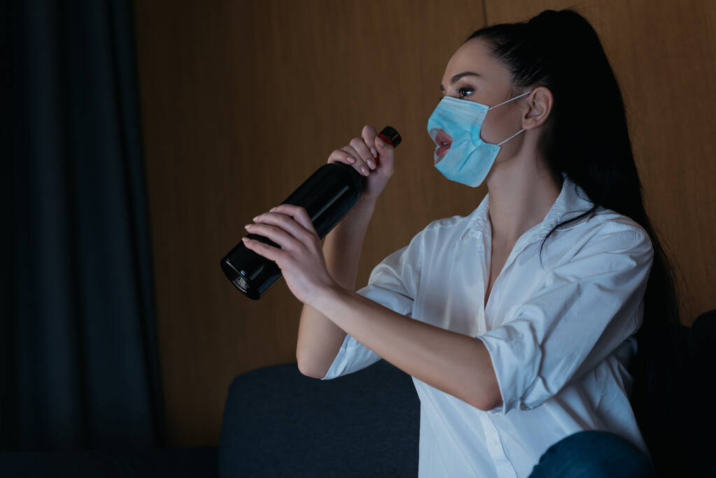 depressed woman in medical mask with hole holding bottle of wine while sitting at home - Photo, Image