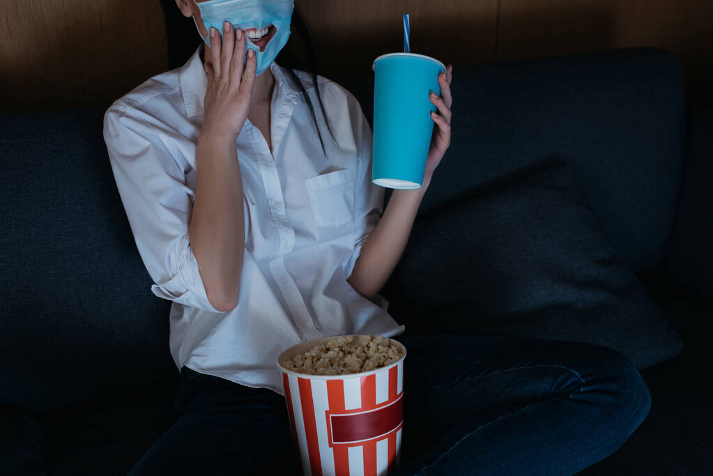 cropped view of smiling woman in medical mask with hole touching face while watching tv with soda and popcorn at home - Photo, Image