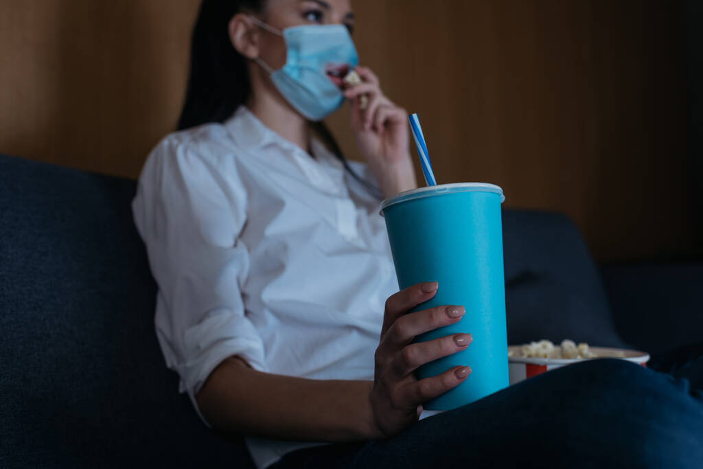 家庭でテレビを見ながらソーダを持ってポップコーンを食べる医療用マスクの若い女性の選択的な焦点 - 写真・画像