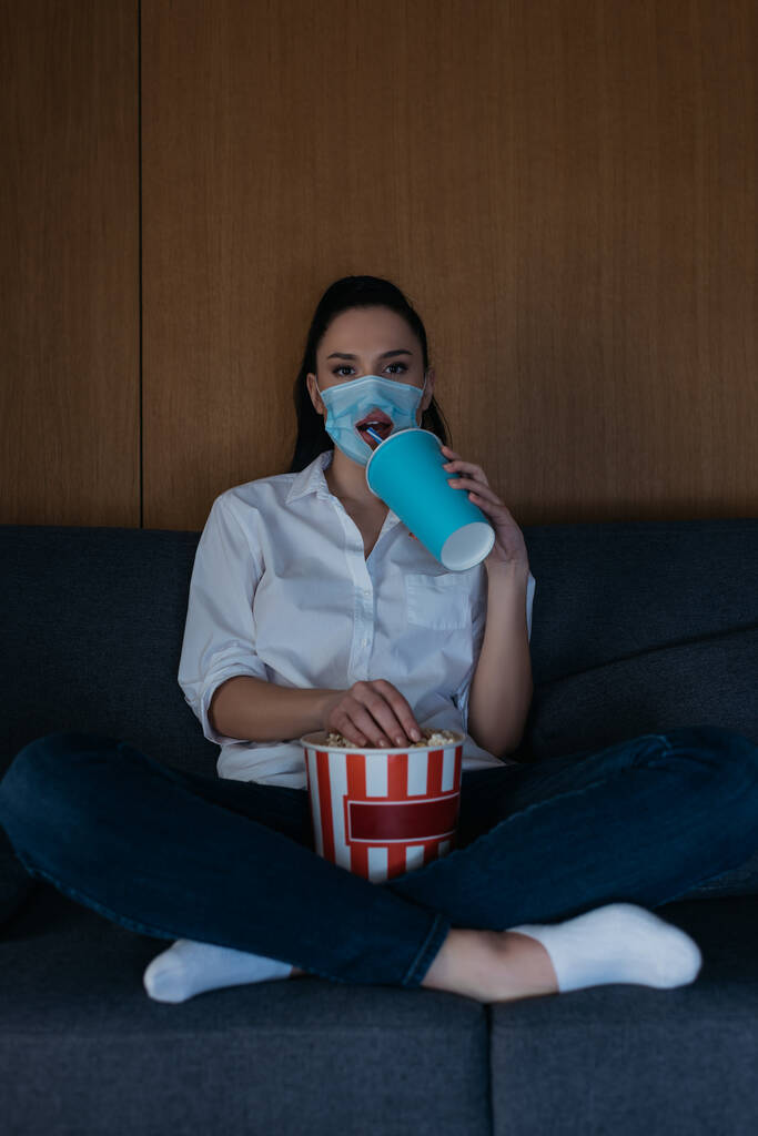 młoda kobieta w masce medycznej z otworem siedzącym na kanapie ze skrzyżowanymi nogami, napojem gazowanym, jedzeniem popcornu i oglądaniem telewizji - Zdjęcie, obraz