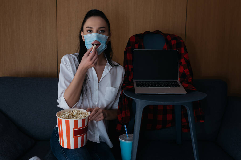 Nuori nainen lääketieteellinen naamio reikä syö popcornia katsellessaan televisiota sohvalla lähellä kannettavaa tietokonetta tyhjällä näytöllä tuolilla
 - Valokuva, kuva