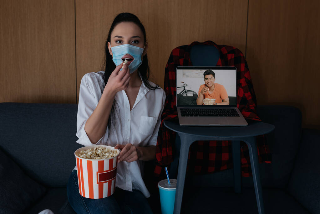 junge Frau in medizinischer Maske mit Loch beim Popcorn essen neben Laptop mit lächelndem asiatischen Freund auf dem Bildschirm - Foto, Bild