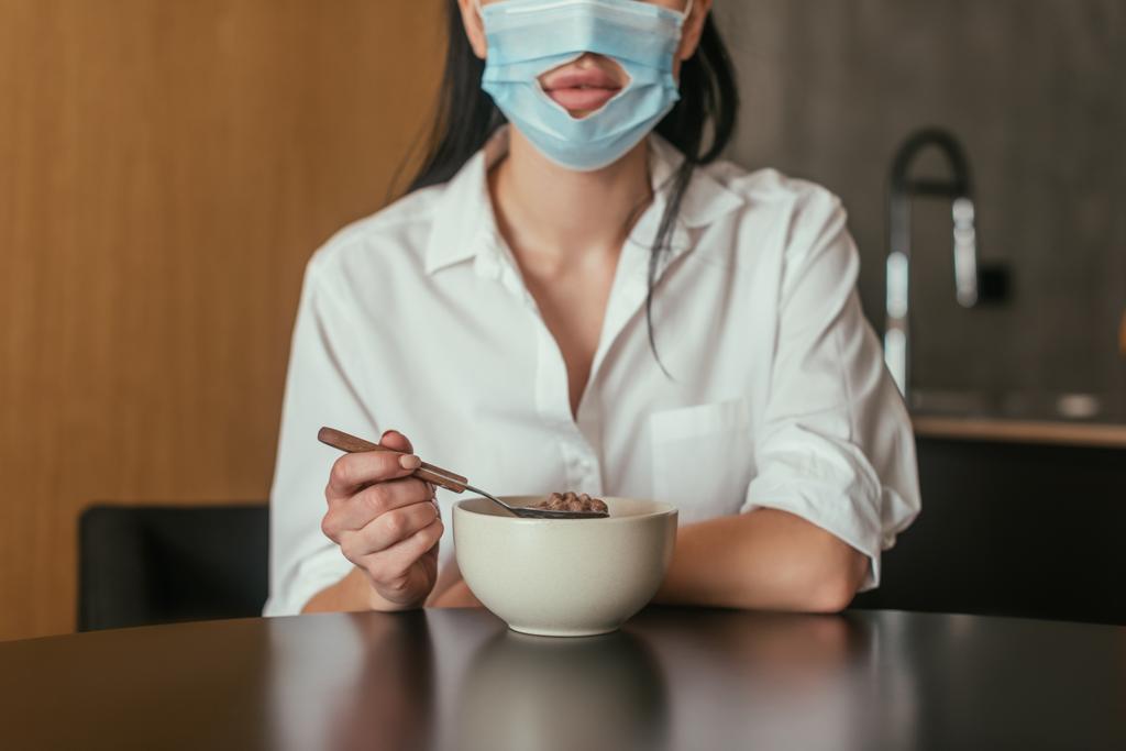 обрезанный вид женщины в медицинской маске с отверстием для рта держа ложку возле миски с завтраком
 - Фото, изображение