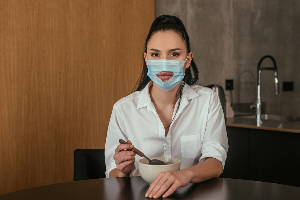 mujer joven seria en máscara médica con agujero mirando a la cámara mientras sostiene la cuchara cerca del tazón con el desayuno
 - Foto, Imagen