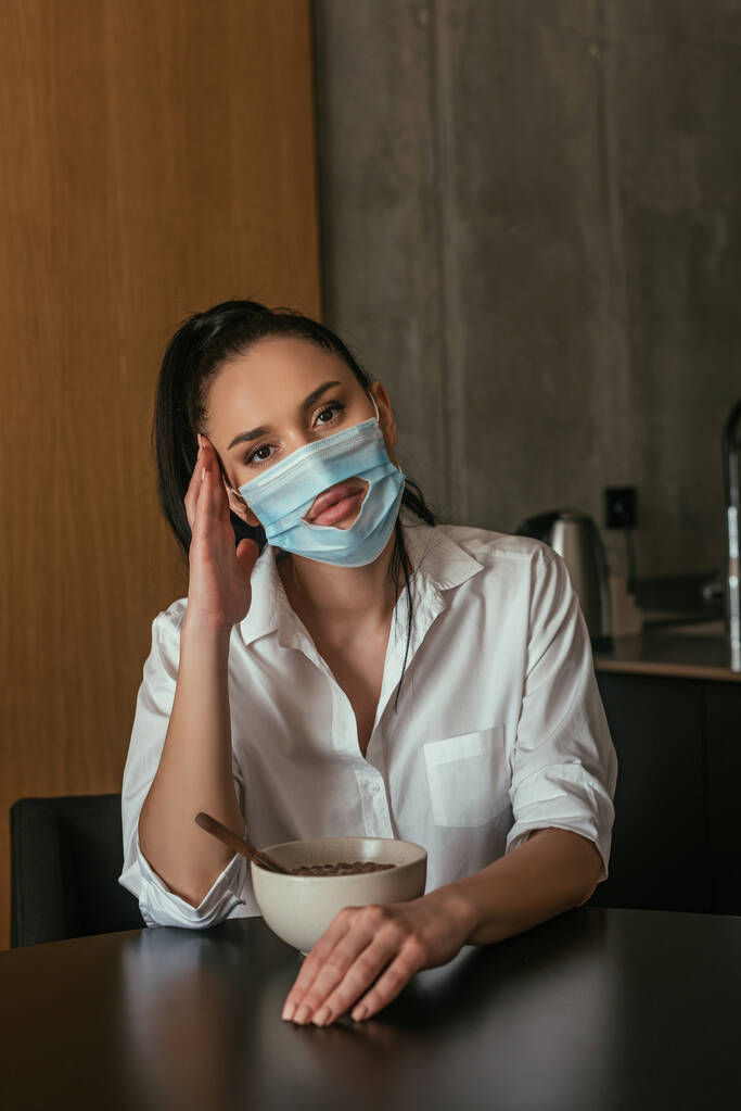 депресивна жінка в медичній масці з отвором, дивлячись на камеру, сидячи біля миски зі сніданком
 - Фото, зображення
