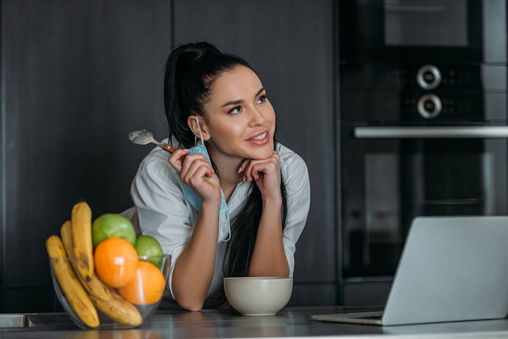 χαμογελαστή, ονειρεμένη γυναίκα χαμογελώντας κρατώντας το κουτάλι κοντά στο laptop, μπολ και φρούτα στην κουζίνα - Φωτογραφία, εικόνα