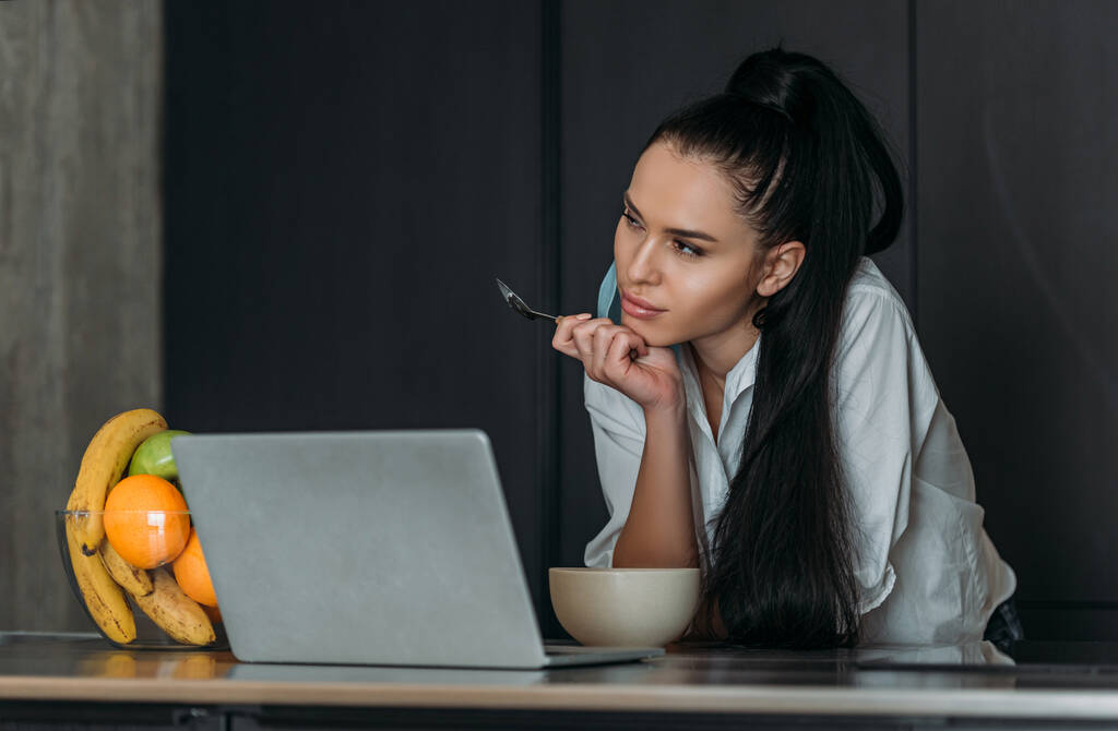 huomaavainen nainen pitää lusikka lähellä kannettavaa tietokonetta, kulhoa ja hedelmiä keittiössä
 - Valokuva, kuva