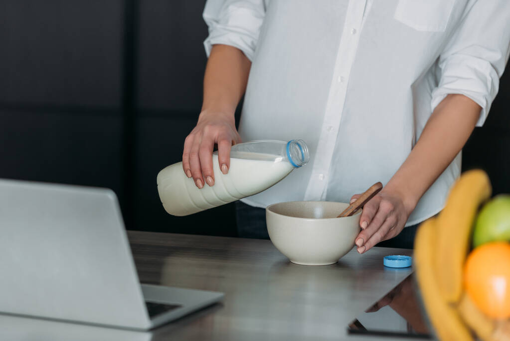 cropped view of woman pouring milk into bowl near laptop in kitchen - Photo, Image