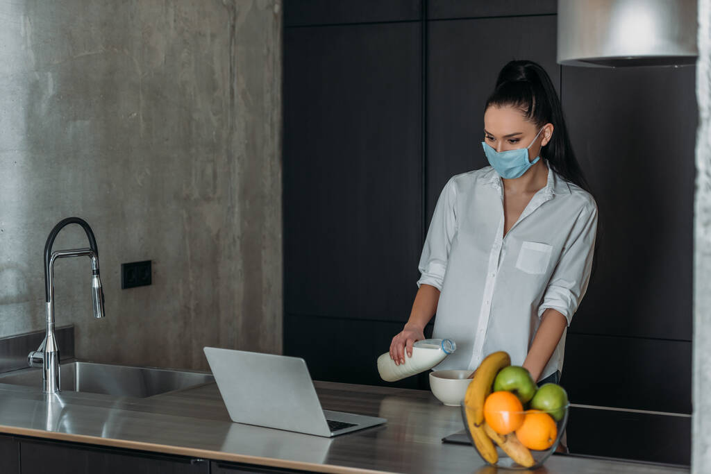 νεαρή γυναίκα με ιατρική μάσκα ρίχνει γάλα στο μπολ, ενώ κοιτάζοντας φορητό υπολογιστή στην κουζίνα - Φωτογραφία, εικόνα
