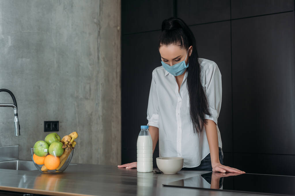 mujer deprimida en máscara médica de pie con la cabeza inclinada cerca de la botella de leche, frutas frescas y tazón en la cocina
 - Foto, Imagen