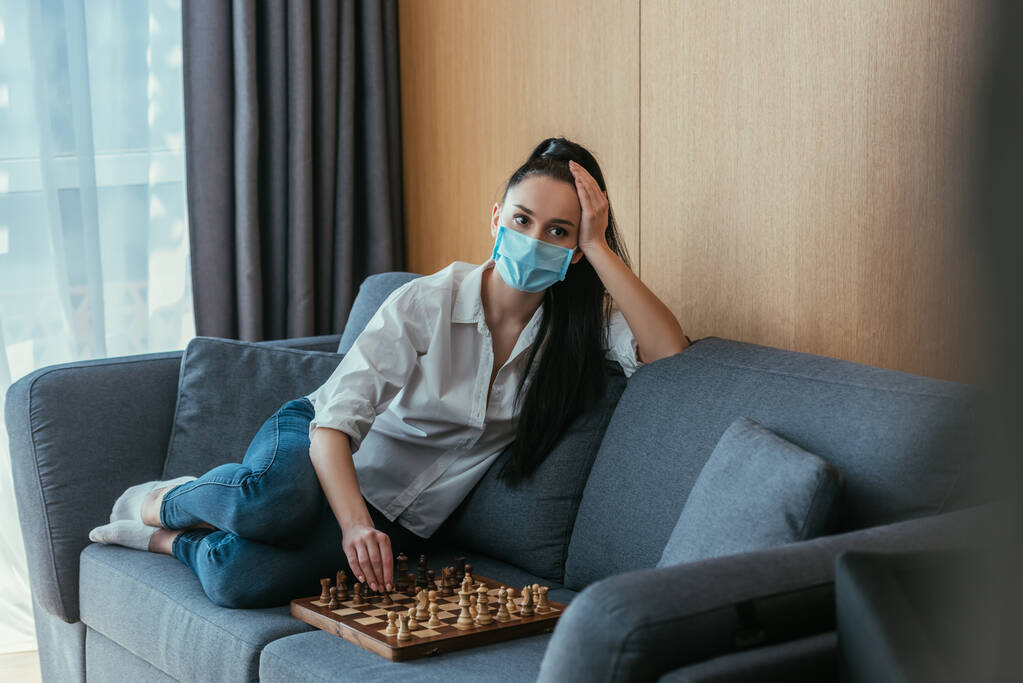 επιλεκτική εστίαση της καταθλιπτικής γυναίκας με προστατευτική μάσκα κάθεται σε καναπέ κοντά σε σκακιέρα  - Φωτογραφία, εικόνα