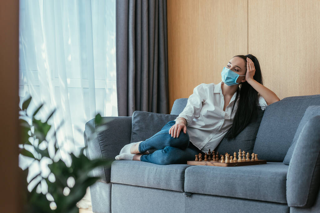 foco seletivo da mulher triste em máscara médica sentado perto do tabuleiro de xadrez e olhando para longe
 - Foto, Imagem