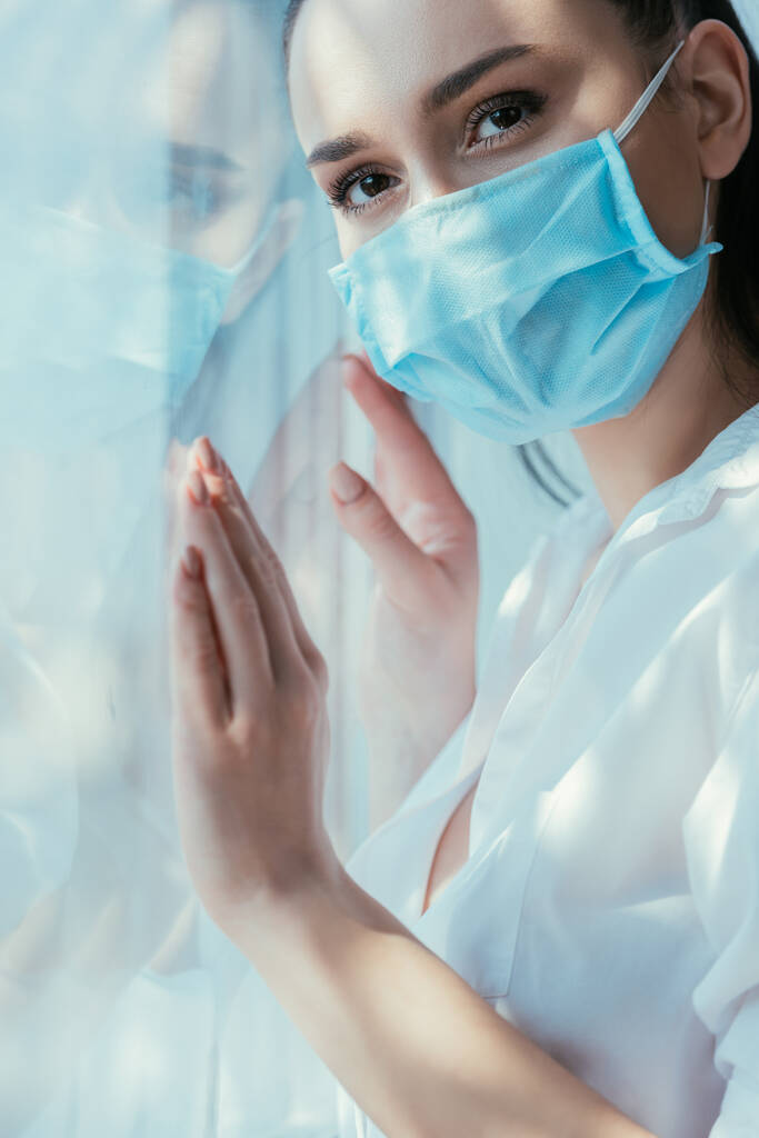 λυπημένο κορίτσι με ιατρική μάσκα αγγίζοντας τζάμι του παραθύρου και κοιτάζοντας μακριά - Φωτογραφία, εικόνα