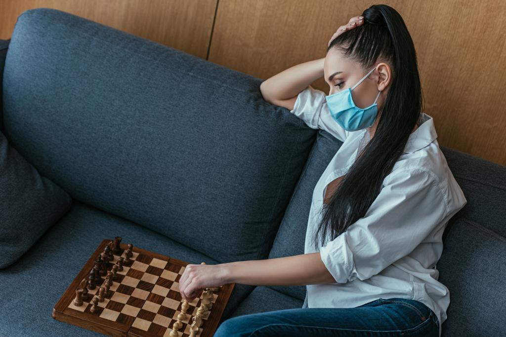 Traurige Frau in medizinischer Maske sitzt auf Sofa neben Schachbrett  - Foto, Bild