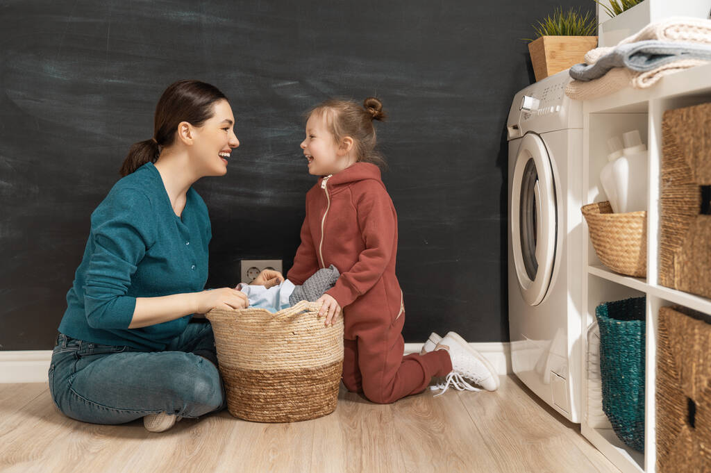 Kaunis nuori nainen ja lapsi tyttö pieni auttaja pitävät hauskaa ja hymyilevät pyykkiä kotona.   - Valokuva, kuva