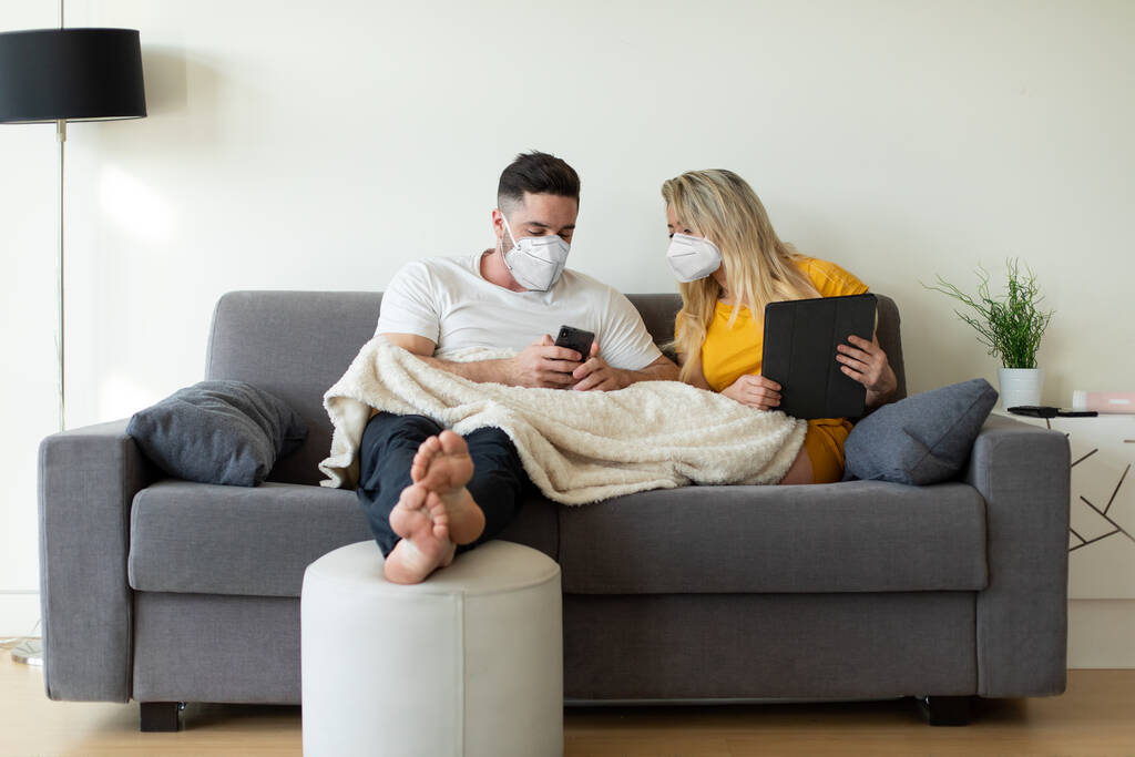Ζευγάρι που φοράει μάσκα προσώπου χρησιμοποιώντας ψηφιακές συσκευές υπό καραντίνα Covid-19. Ψηφιακή αναψυχή στο σπίτι υπό coronavirus παγκόσμια κρίση, - Φωτογραφία, εικόνα