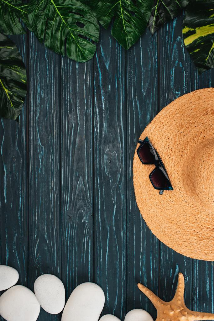 Bovenaanzicht van stro hoed en zonnebril in de buurt van bladeren en zee stenen op donkere houten achtergrond - Foto, afbeelding