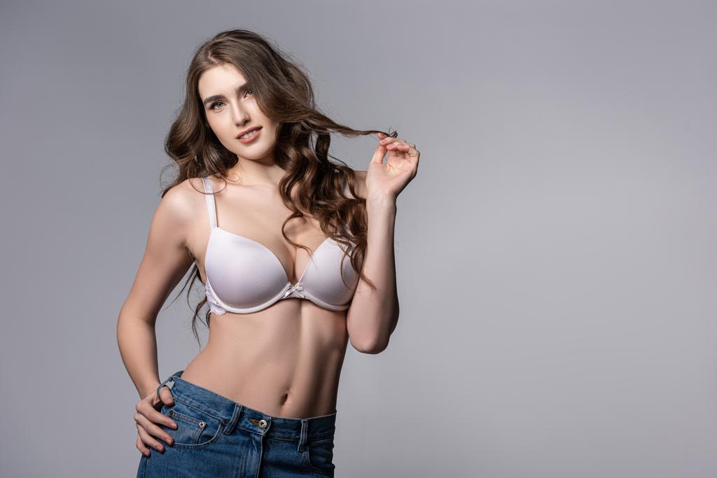 attractive girl in bra and jeans standing and touching hair isolated on grey  - Photo, Image