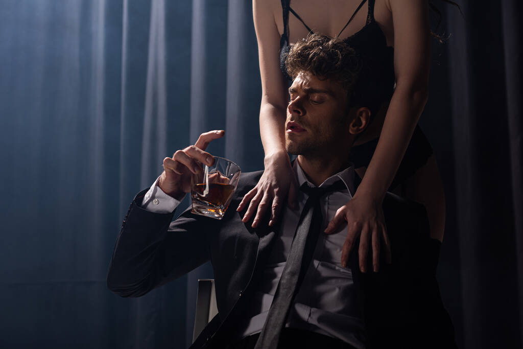 σαγηνευτική γυναίκα αγγίζει τον άνδρα κάθεται και κρατώντας ένα ποτήρι ουίσκι σε μαύρο - Φωτογραφία, εικόνα