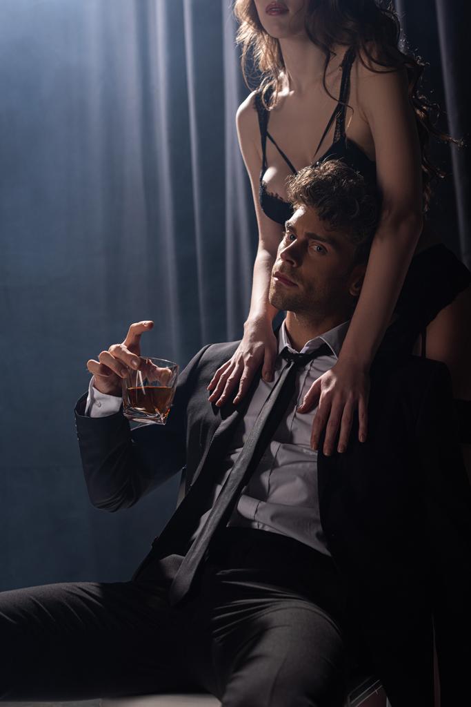 соблазнительная женщина прикасается к красивому мужчине сидя и держа стакан виски на черном
 - Фото, изображение